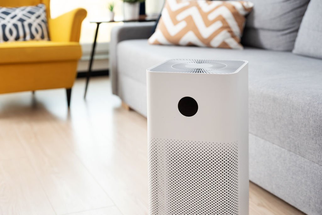 Contemporary white air purifier on a wooden floor, with a vibrant yellow armchair and patterned cushions in the background, illustrating how to use an air purifier in home decor