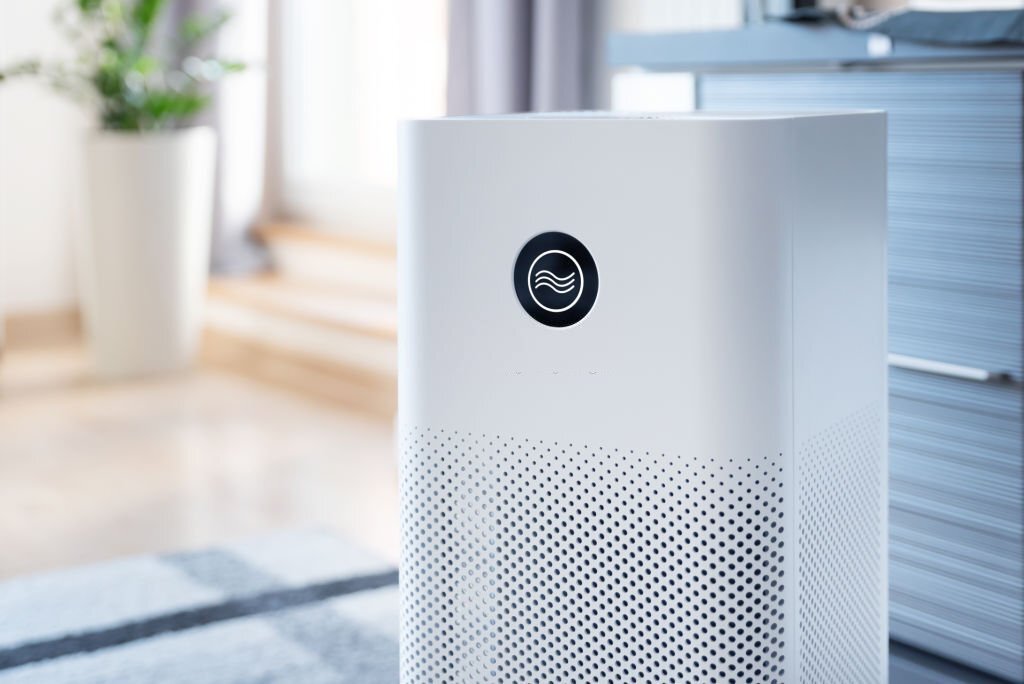 Modern white air purifier with a simple design and perforated lower half, possibly utilizing a charcoal filter, set in a bright room with plants