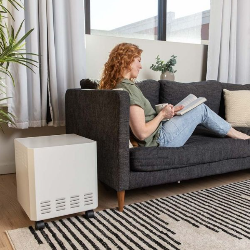 White EnviroKlenz Mobile Air System near a dark sofa with a woman reading, enhancing air quality in a cozy living room setting