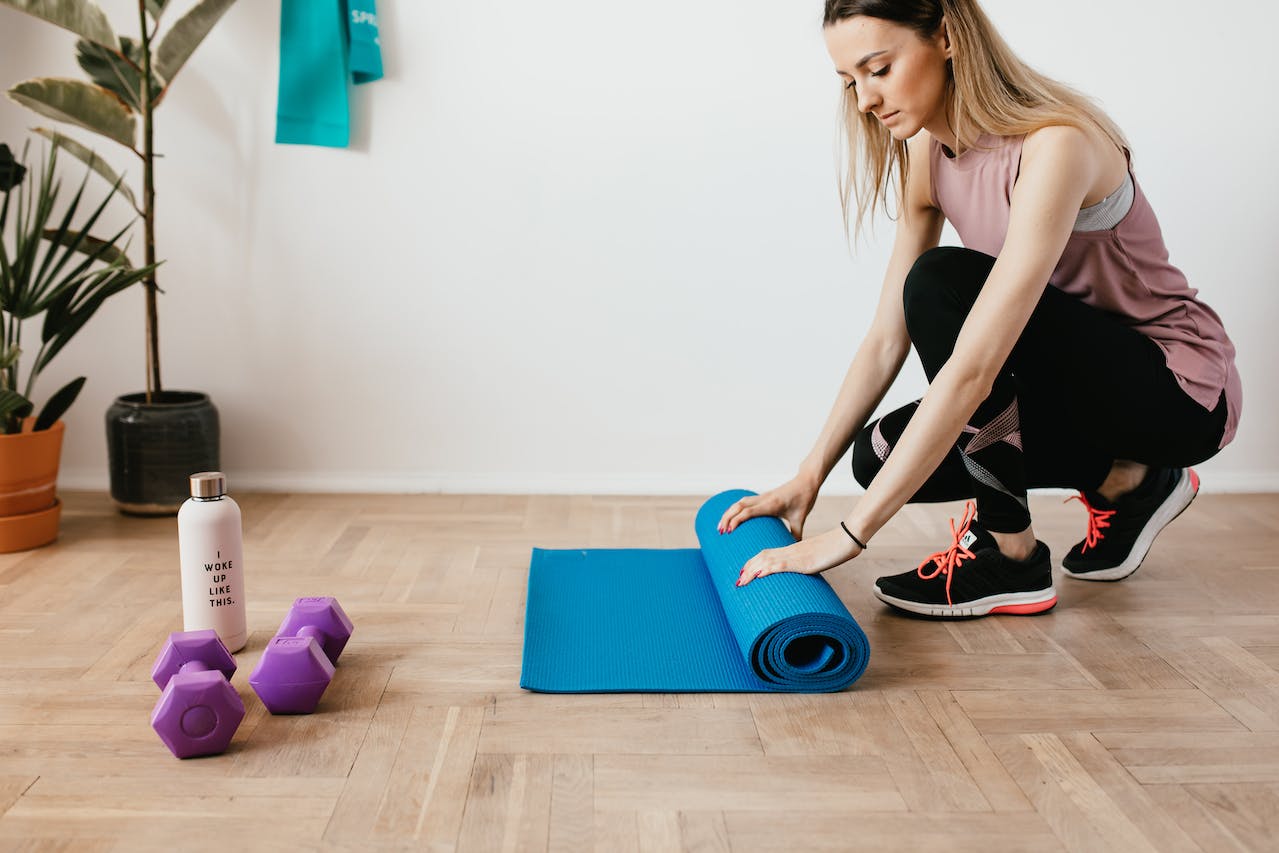 Woman unrolling a blue yoga mat in a well-ventilated home gym space with a portable air purifier, dumbbells, and a water bottle, promoting a clean and healthy exercise environment.