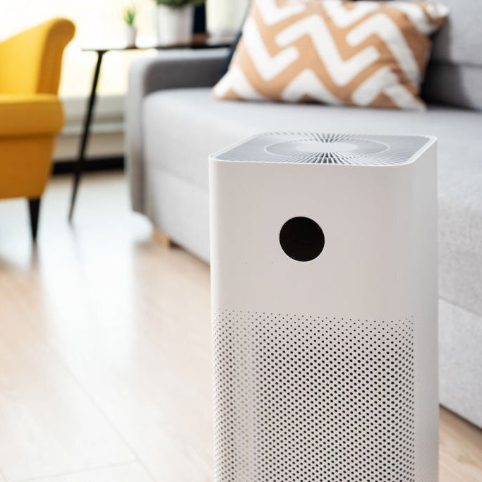 Contemporary white air purifier on a wooden floor, with a vibrant yellow armchair and patterned cushions in the background, illustrating how to use an air purifier in home decor